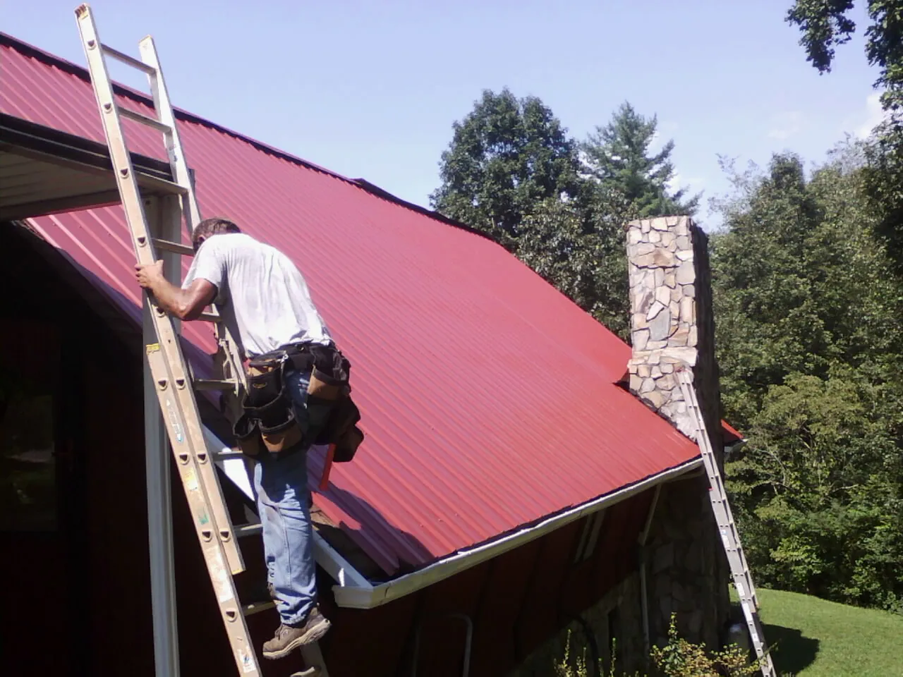 Roofer on a ladder beside a red metal roof and stone chimney — Soffit & Fascia Repair by Roofing Brothers in Las Cruces